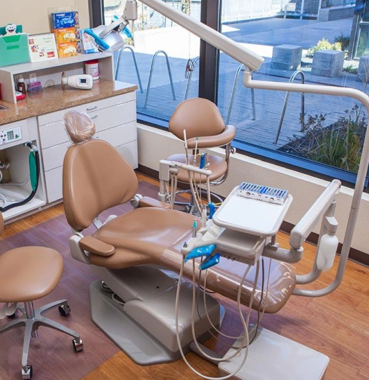 Modern dental operatory with tan patient chair, overhead light, and equipment in bright clinical setting.