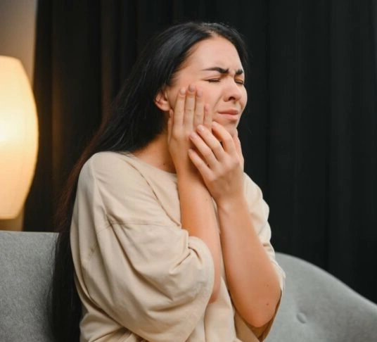 Woman experiencing tooth pain, holding cheek, seated indoors with warm lighting.