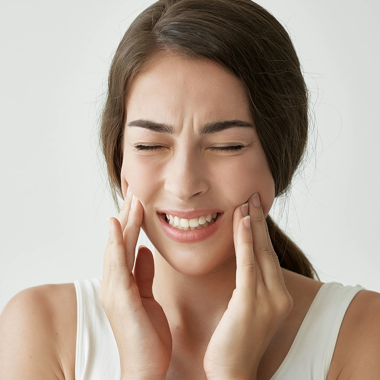 Woman with tooth pain holding cheeks, smiling against white background.