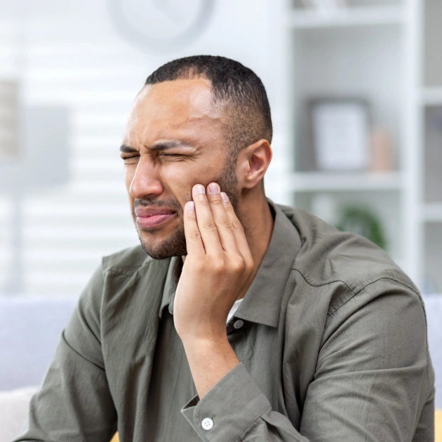 Man in olive shirt touching jaw in discomfort, modern dental office background.
