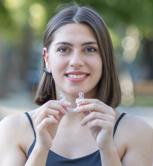Young woman holding clear aligner, smiling outdoors with blurred green background.