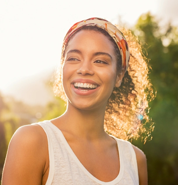 Young woman smiling outdoors in sunlight wearing white tank top and headband.