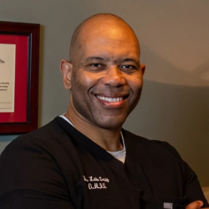 Headshot of a smiling, bald Black man in black scrubs, featuring a slightly darker, high-contrast exposure that deepens the shadows on the background wall.