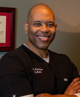 Professional headshot of a smiling, bald Black man in black medical scrubs, featuring a slightly cooler color tone and tighter top-and-bottom cropping